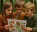 Drei Kinder betrachten im Wald eine bunte Zeichnung mit Tieren des Waldes – darunter Fuchs, Eule, Reh und Frosch – und rätseln gemeinsam mit Lupe und Neugier